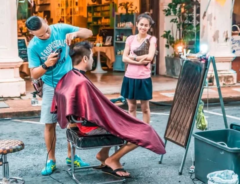 Парикмахер стрижёт клиента на улице, рядом наблюдает девушка.