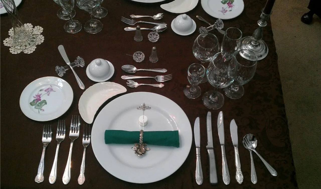 Изысканная сервировка стола с белой посудой, серебряными столовыми приборами и хрустальной посудой на фоне темной скатерти.